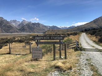 A rural landscape featuring a gravel road leading towards a mountainous horizon. The foreground has a sign reading 'Erewhon Station' and 'Home of the Working Horse', and a directional sign indicating a turning area 2 km ahead. The area is surrounded by dry grasslands with tall mountains in the background under a clear blue sky.