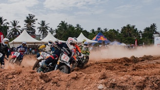 A group of motocross riders navigating through a dirt track with intense speed and agility. The scene is set outdoors with palm trees and white tents in the background, while dust clouds are kicked up by the motorcycles.