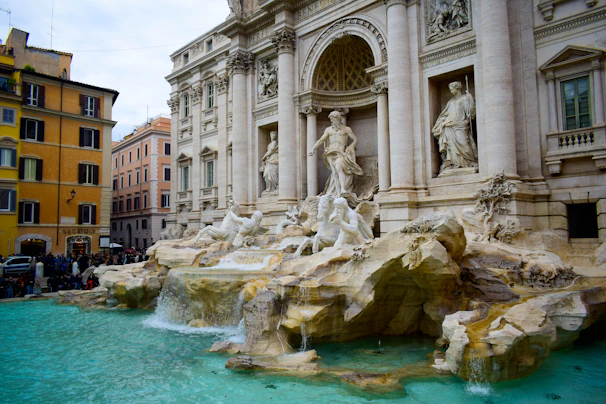 A large, ornate baroque fountain with intricate stone sculptures is situated in a bustling urban plaza. The architecture features classical elements with columns and statues depicting mythological figures, surrounded by cascading water. Adjacent to the fountain, colorful historic buildings line the street, adding to the scenic ambiance.