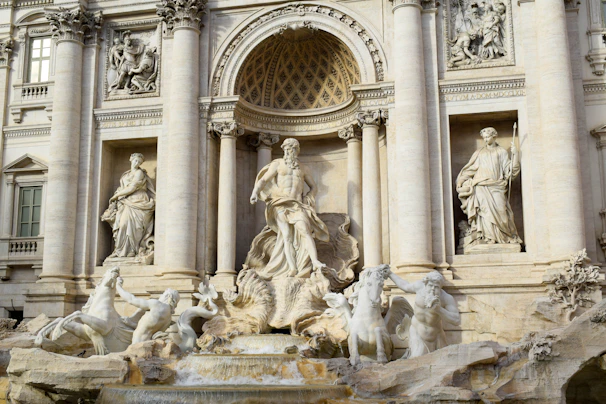 A grand and ornate Baroque fountain, featuring a central statue of a male figure surrounded by other smaller statues and horses. The fountain is elaborately carved from white stone and integrated into a building façade with columns and relief sculptures. Water flows dramatically from the base.