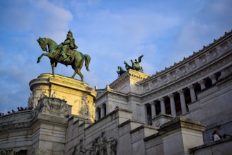 A grand architectural structure features a large equestrian statue of a historical figure atop a pedestal. The monument is adorned with intricate bas-reliefs and classical columns, accented by statues of winged horses. The sky is partly cloudy, casting a warm light on the stone structure.