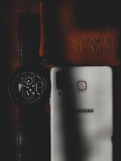 A wristwatch with a dark strap and round dial is placed next to a white smartphone that has the brand name embossed on it. There is also a leather item, likely a wallet or notebook, embossed with the words 'Genuine Leather'. The scene is dimly lit, with shadows creating a moody aesthetic.
