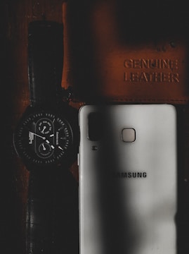 A wristwatch with a dark strap and round dial is placed next to a white smartphone that has the brand name embossed on it. There is also a leather item, likely a wallet or notebook, embossed with the words 'Genuine Leather'. The scene is dimly lit, with shadows creating a moody aesthetic.