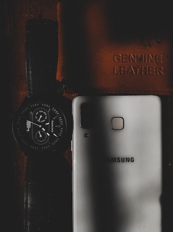 A wristwatch with a dark strap and round dial is placed next to a white smartphone that has the brand name embossed on it. There is also a leather item, likely a wallet or notebook, embossed with the words 'Genuine Leather'. The scene is dimly lit, with shadows creating a moody aesthetic.