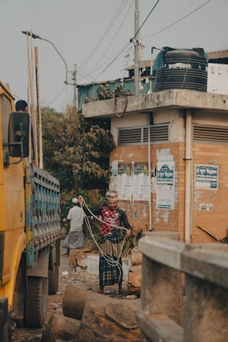 A man wearing a patterned shirt and a traditional headscarf is handling a rope in a street environment. Nearby, a large yellow truck is parked next to a brick building adorned with various posters. Other people are visible in the background, walking along a debris-strewn path. Utility poles and wires tower above the scene, and logs are scattered in the foreground.