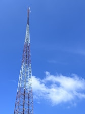 A tall communication tower with a lattice structure reaching into a clear blue sky. The tower is composed of metal beams and features small antennas near the top. A few white clouds are visible, enhancing the sense of height and openness.