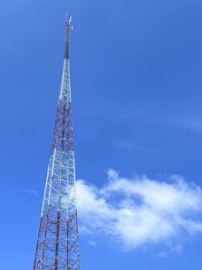 A tall communication tower with a lattice structure reaching into a clear blue sky. The tower is composed of metal beams and features small antennas near the top. A few white clouds are visible, enhancing the sense of height and openness.
