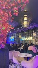 The scene features a lively restaurant interior with several people gathered around a table, enjoying a social event. A decorative ceiling adorned with vibrant red and purple flowers complements the modern light fixtures. The patterned black and white floor and contemporary wall design add to the stylish ambiance. The word 'SAPORES' is visible on the wall.