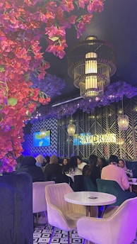 The scene features a lively restaurant interior with several people gathered around a table, enjoying a social event. A decorative ceiling adorned with vibrant red and purple flowers complements the modern light fixtures. The patterned black and white floor and contemporary wall design add to the stylish ambiance. The word 'SAPORES' is visible on the wall.