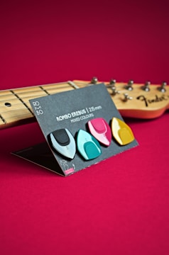 A set of colorful guitar picks displayed on a stand, with a guitar neck and tuning pegs visible in the background. The guitar picks are arranged in a manner that showcases their shapes and colors, set against a rich red backdrop.