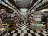 A narrow aisle of a bookshop with floor-to-ceiling shelves filled with a variety of books and magazines. The space has a checkerboard black and white tile floor, and the books are organized in neat rows with numerous covers visible. The shelving stretches far into the background, creating a sense of depth and abundance of reading material.