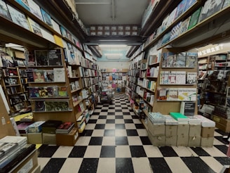A narrow aisle of a bookshop with floor-to-ceiling shelves filled with a variety of books and magazines. The space has a checkerboard black and white tile floor, and the books are organized in neat rows with numerous covers visible. The shelving stretches far into the background, creating a sense of depth and abundance of reading material.