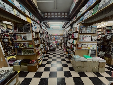 A narrow aisle of a bookshop with floor-to-ceiling shelves filled with a variety of books and magazines. The space has a checkerboard black and white tile floor, and the books are organized in neat rows with numerous covers visible. The shelving stretches far into the background, creating a sense of depth and abundance of reading material.
