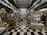 A narrow aisle of a bookshop with floor-to-ceiling shelves filled with a variety of books and magazines. The space has a checkerboard black and white tile floor, and the books are organized in neat rows with numerous covers visible. The shelving stretches far into the background, creating a sense of depth and abundance of reading material.