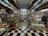 A narrow aisle of a bookshop with floor-to-ceiling shelves filled with a variety of books and magazines. The space has a checkerboard black and white tile floor, and the books are organized in neat rows with numerous covers visible. The shelving stretches far into the background, creating a sense of depth and abundance of reading material.