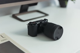A digital camera with a large lens and a prominent brand logo on its body is placed on a modern white desk. In the background, a monitor stand and part of a monitor are visible. A magazine or book with a minimalistic cover lies nearby, and a small plant is partially visible, adding a touch of nature to the scene.