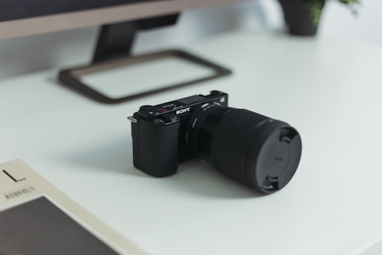 A digital camera with a large lens and a prominent brand logo on its body is placed on a modern white desk. In the background, a monitor stand and part of a monitor are visible. A magazine or book with a minimalistic cover lies nearby, and a small plant is partially visible, adding a touch of nature to the scene.