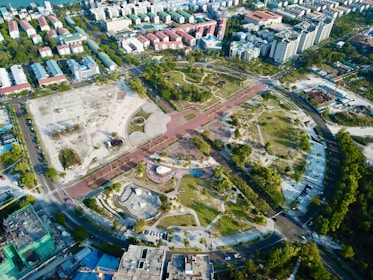The aerial view showcases a large, semi-circular urban park surrounded by roads and buildings. The park features various landscaped sections with green trees, pathways, and open grassy areas. To the left, there is a section with construction or clear land. Surrounding the park are numerous residential and commercial buildings of varying sizes. The arrangement suggests a well-planned city area with optimal use of space.