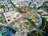 The aerial view showcases a large, semi-circular urban park surrounded by roads and buildings. The park features various landscaped sections with green trees, pathways, and open grassy areas. To the left, there is a section with construction or clear land. Surrounding the park are numerous residential and commercial buildings of varying sizes. The arrangement suggests a well-planned city area with optimal use of space.