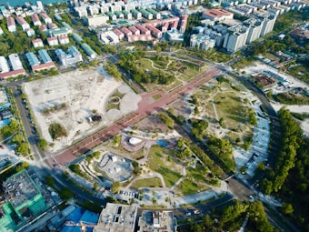 The aerial view showcases a large, semi-circular urban park surrounded by roads and buildings. The park features various landscaped sections with green trees, pathways, and open grassy areas. To the left, there is a section with construction or clear land. Surrounding the park are numerous residential and commercial buildings of varying sizes. The arrangement suggests a well-planned city area with optimal use of space.