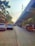 A busy urban street is shown with parked cars on the left, auto rickshaws, and motorcycles moving on the road. A large elevated railway or metro track runs parallel to the road. Trees and buildings line the street, with some people visible on the sidewalk.