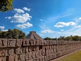A well-preserved ancient pyramid, surrounded by lush green foliage under a bright, sunny sky. The foreground features a stone wall intricately carved with faces, symbolizing historical or cultural significance.