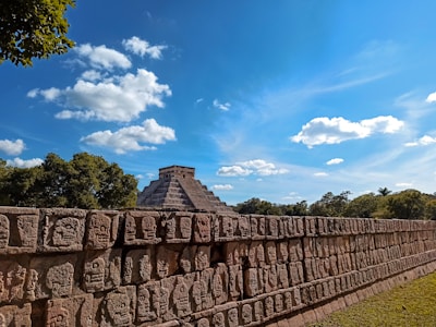 A well-preserved ancient pyramid, surrounded by lush green foliage under a bright, sunny sky. The foreground features a stone wall intricately carved with faces, symbolizing historical or cultural significance.
