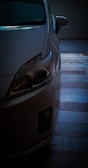 A close-up of the front left corner of a car, partially in shadow. The headlight is prominently featured, and the background includes a floor with a striped pattern.