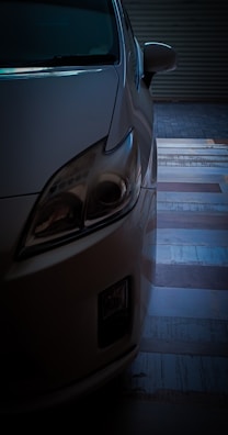 A close-up of the front left corner of a car, partially in shadow. The headlight is prominently featured, and the background includes a floor with a striped pattern.