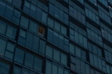 A grid of reflective glass windows covers the facade of a modern high-rise building. Most windows are dark, but one is lit with a warm glow, suggesting interior lighting.