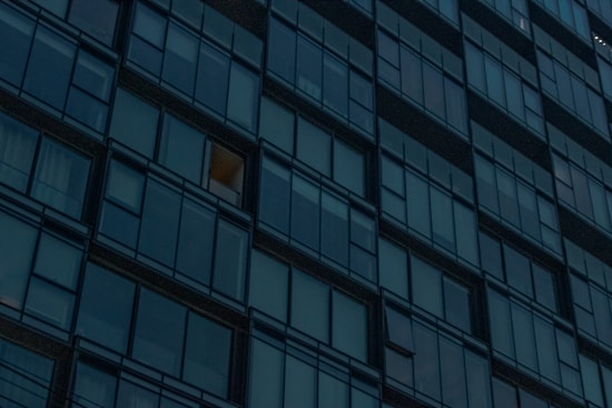 A grid of reflective glass windows covers the facade of a modern high-rise building. Most windows are dark, but one is lit with a warm glow, suggesting interior lighting.