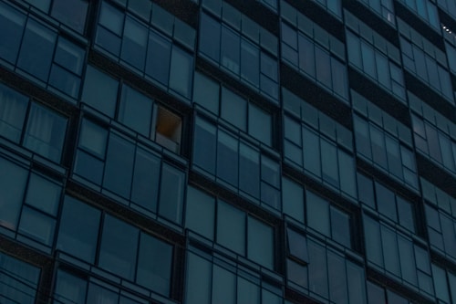 A grid of reflective glass windows covers the facade of a modern high-rise building. Most windows are dark, but one is lit with a warm glow, suggesting interior lighting.