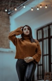 A person wearing a brown knit sweater and black leather pants stands confidently in an outdoor setting. The background features soft-focus urban architecture and string lights that add a decorative touch.
