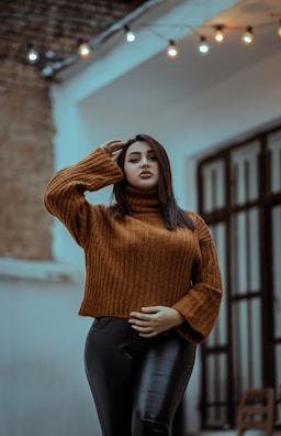 A person wearing a brown knit sweater and black leather pants stands confidently in an outdoor setting. The background features soft-focus urban architecture and string lights that add a decorative touch.