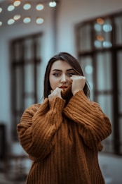 A person is wrapped in a large, cozy brown sweater. They are holding the collar up close to their face, partially obscuring their mouth. The background is softly blurred, with bokeh lights in the distance and large windows in view.