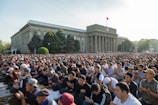 A large crowd of people gathered outside a government building engaged in prayer or meditation. The building features classical architecture with columns. Trees and greenery surround the area, and a flag is seen atop the structure.