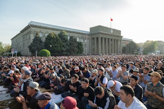 A large crowd of people gathered outside a government building engaged in prayer or meditation. The building features classical architecture with columns. Trees and greenery surround the area, and a flag is seen atop the structure.