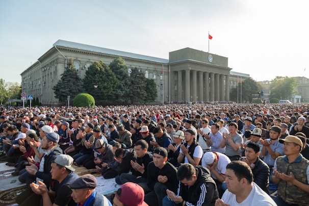 A large crowd of people gathered outside a government building engaged in prayer or meditation. The building features classical architecture with columns. Trees and greenery surround the area, and a flag is seen atop the structure.