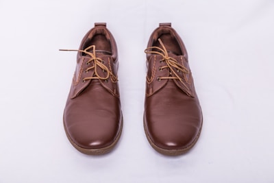 A pair of brown leather shoes with laces, placed side by side on a light gray background. The shoes have a polished finish and feature detailed stitching along the seams.