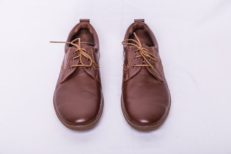 A pair of brown leather shoes with laces, placed side by side on a light gray background. The shoes have a polished finish and feature detailed stitching along the seams.