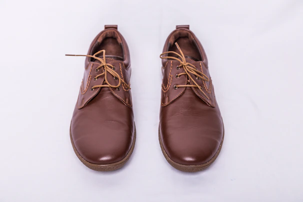 A pair of brown leather shoes with laces, placed side by side on a light gray background. The shoes have a polished finish and feature detailed stitching along the seams.