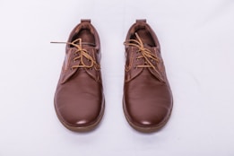 A pair of brown leather shoes with laces, placed side by side on a light gray background. The shoes have a polished finish and feature detailed stitching along the seams.