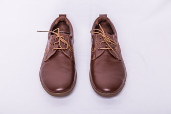 A pair of brown leather shoes with laces, placed side by side on a light gray background. The shoes have a polished finish and feature detailed stitching along the seams.