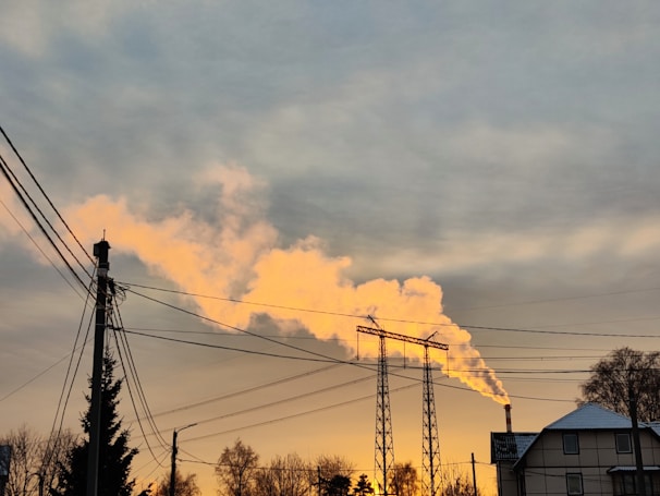 A sunset sky with industrial smoke rising from a factory chimney, casting an orange glow. Power lines and a residential building are visible in the foreground, along with silhouettes of trees.