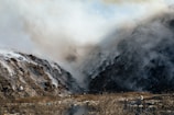 Dense smoke billows between two large piles of debris and waste, creating a hazy atmosphere. The ground is uneven with visible dry vegetation and scattered litter. The scene is dominated by shades of gray from the smoke and the dark tones of the waste piles.