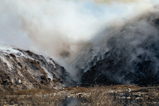 Dense smoke billows between two large piles of debris and waste, creating a hazy atmosphere. The ground is uneven with visible dry vegetation and scattered litter. The scene is dominated by shades of gray from the smoke and the dark tones of the waste piles.