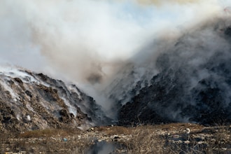 Dense smoke billows between two large piles of debris and waste, creating a hazy atmosphere. The ground is uneven with visible dry vegetation and scattered litter. The scene is dominated by shades of gray from the smoke and the dark tones of the waste piles.