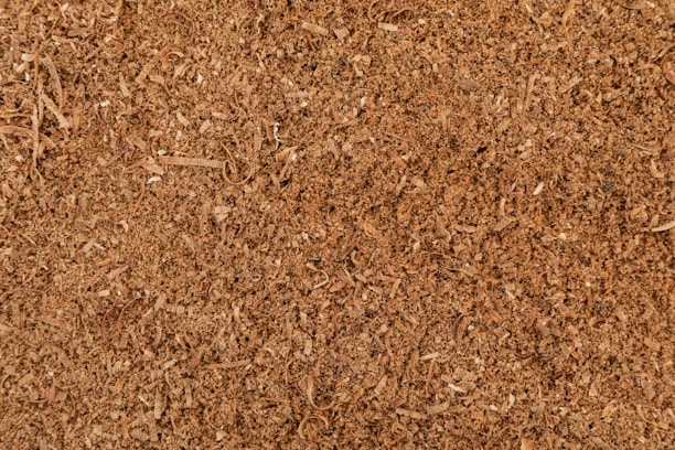 A close-up view of a textured surface composed of small wood shavings and sawdust. The surface appears uneven, with varying shades of brown and visible particles of wood fibers.