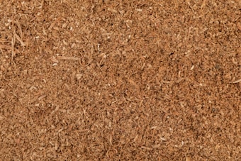 A close-up view of a textured surface composed of small wood shavings and sawdust. The surface appears uneven, with varying shades of brown and visible particles of wood fibers.