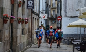 A group of hikers with backpacks and outdoor gear walks down a narrow cobblestone street between old stone buildings. Brightly colored flowers in pots are mounted on the wall, and there are several restaurants and cafes with outdoor seating areas featuring large umbrellas.